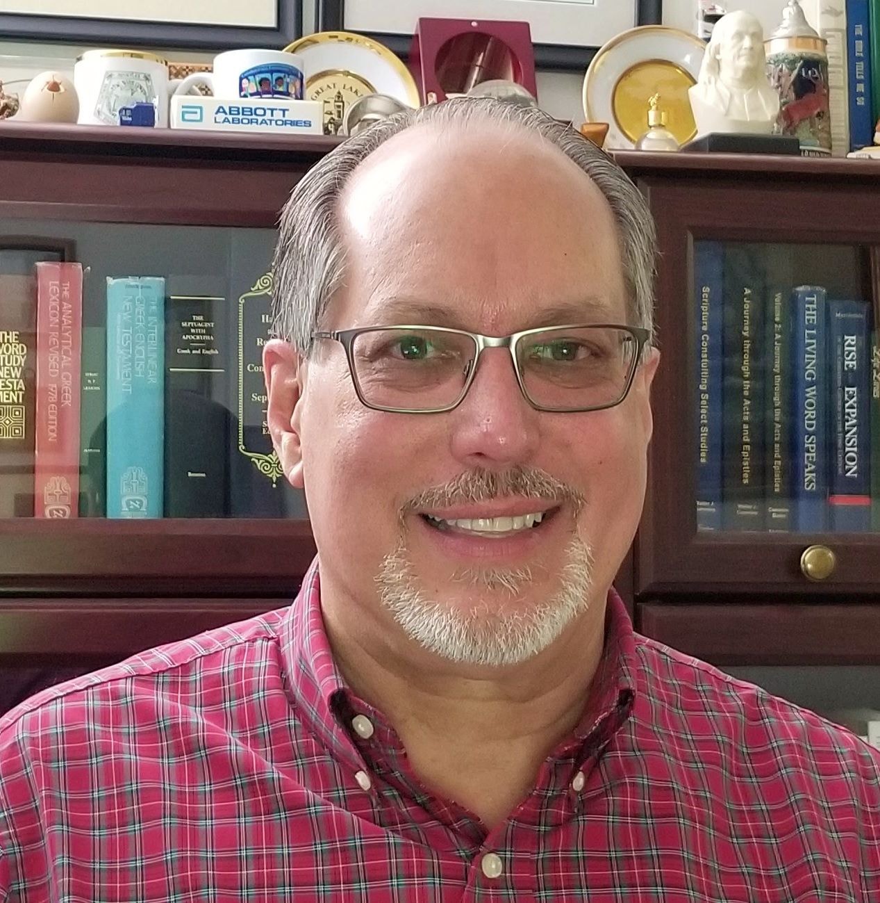 Smiling man with glasses and a red plaid shirt in front of a bookshelf.