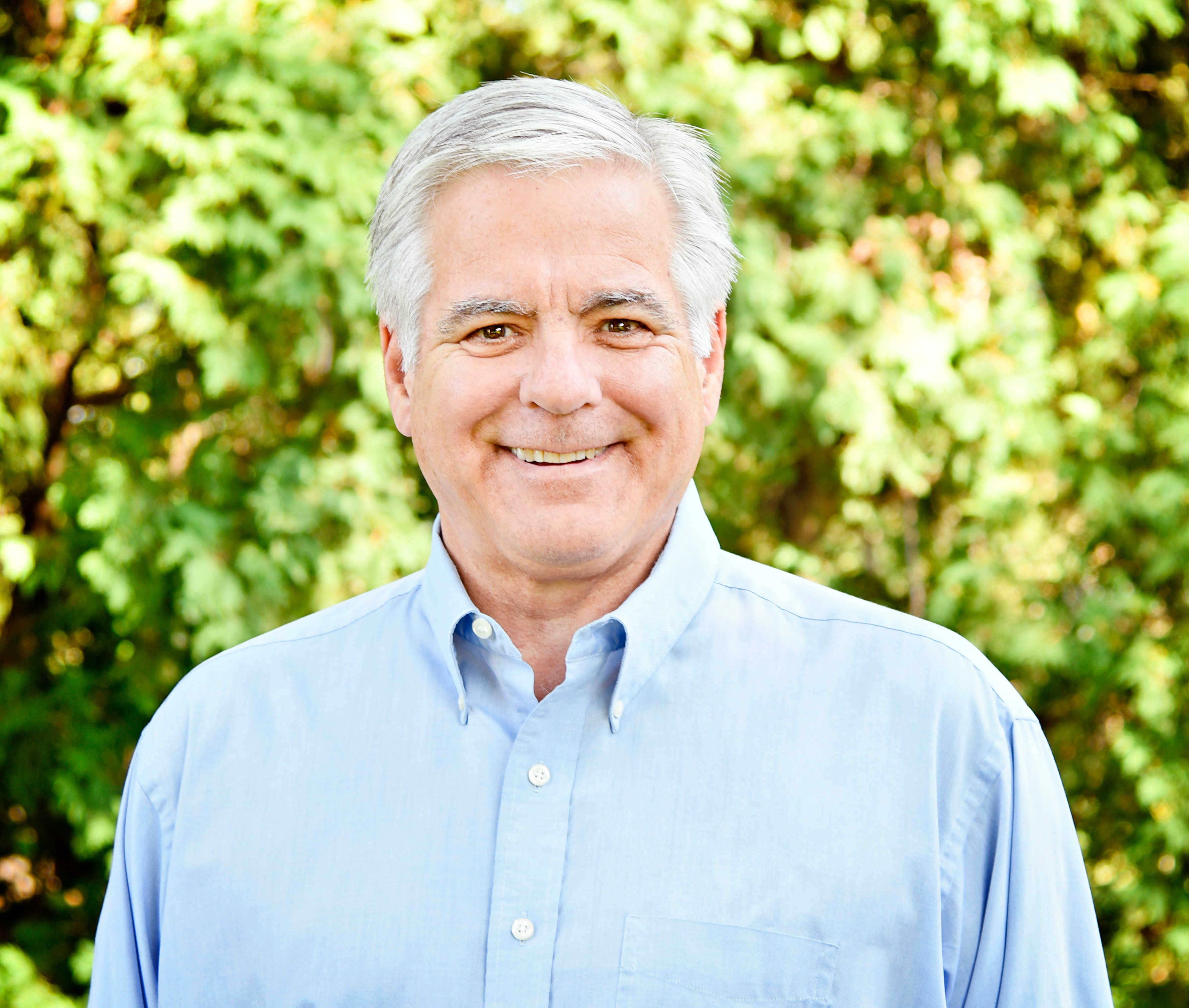 Smiling elderly man outdoors with greenery background.